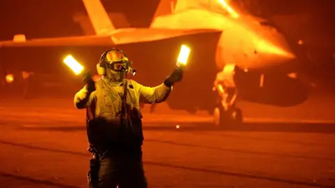 Deck crew member holds up luminous sticks to guide jets around the flight deck at night