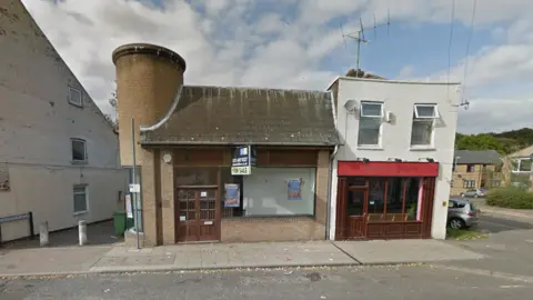 Google Single-storey building with round brick tower to the left. The front of the building has a large wooden door and picture window, which has a white board on the inside.