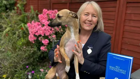 Dawn Murray A woman with shoulder-length grey hair is holding a whippet-style dog. She is wearing a smart navy jacket over a white top.