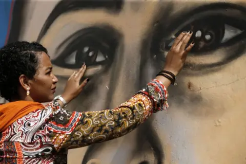 AFP Sudanese activist Eythar Gubara holds her hands up to the eyes of a large, painted face on a wall. Her expression is thoughtful.