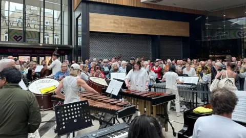 People playing instruments in the Broadway shopping centre with people watching.