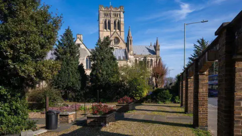Watsons A cathedral building is shown in the background of a front yard featuring paving slabs, flowers beds and trees.