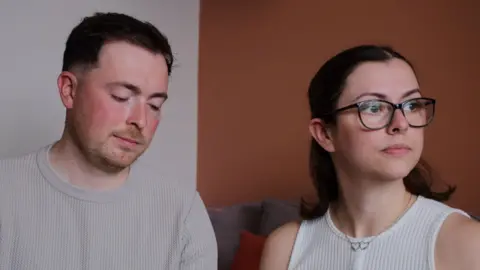 The McCreadys are sitting together on a sofa. Lauryn is looking wistfully out of a window while Andrew is looking downards towards their daughter's memory box