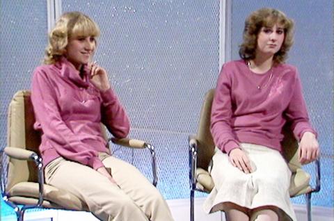 Both twins sitting in the studio. They are both wearing pink jumpers, one with cream trousers and the other with a white skirt.