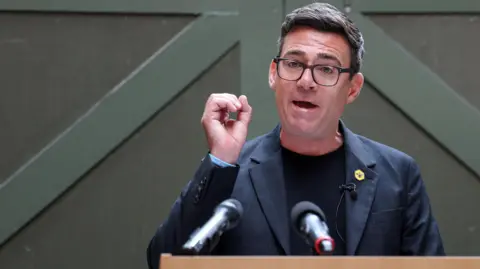 Andy Burnham, making a speech, wearing a dark T-shirt and grey jacket with a badge of a bee on the lapel. 