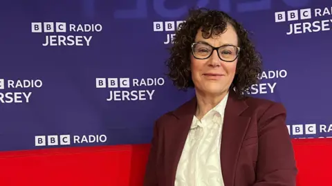 Carmel is is seated on a bright red curved sofa in front of a backdrop that reads “BBC Radio Jersey” repeatedly in white text on a blue background. She is dressed in a maroon suit with a white shirt and is wearing a wristwatch. The setting suggests a media or interview environment.