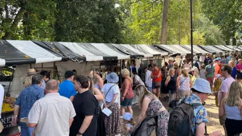 A crowd of people stood near black and white market stalls, with trees surrounding the area.