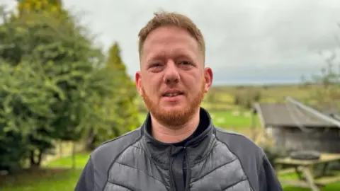 Nick Spann Man with ginger beard standing in front of trees