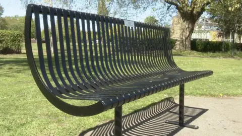 John Kinder The photo shows a black metal bench which is in a park. It has a silver plaque on it. 