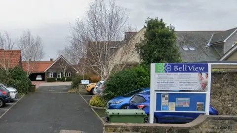 Google A sign saying Bell View is in the foreground of the picture and behind you can see a modern one storey building. There is a driveway in front with parking spaces in either side  