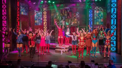 About two dozen people, in costumes and on stage, have their arms raised as they perform the Kinky Boots production. They are all wearing thigh-high boots of different colours and designs, and the stage is lit with other shoe designs.