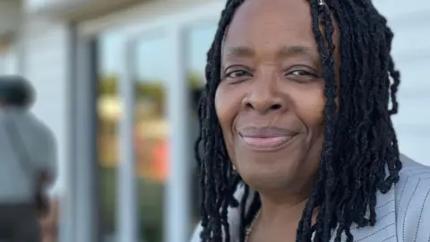 Jacqui Wilson smiles into the camera. She has black braids and is wearing a grey jacket. 