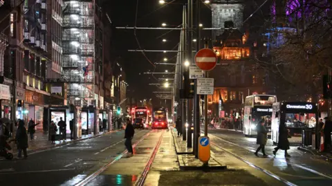 PA Media A view looking down Princes Street, with buses and trams travelling. The street is quiet with only a few people walking around, the sky is clear and dark, but dry.