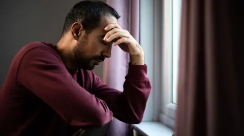 Getty Images Man in maroon jumper with his head in his hands, sat by his window. He has dark hair and dark facial hair.