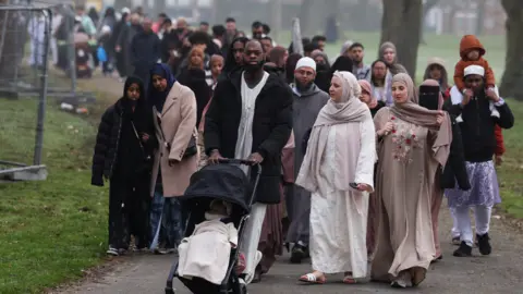Reuters Muslim women dressed in beige abayas and a man in a white thawb pushing a black pram with a small child girl wrapped in a white blanket. They are arriving in a park in Birmingham.