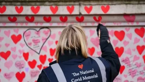 PA Media A volunteer from the Covid-19 Bereaved Families for Justice campaign group paints a heart on the National Covid Memorial Wall opposite the Palace of Westminster in central London,