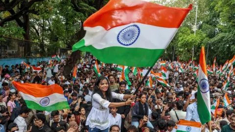 A woman waves a large Indian flag in the midst of a crowd carrying similar flags. She wears a white blouse and blue jeans, and is smiling broadly. Trees surround the large crowd.