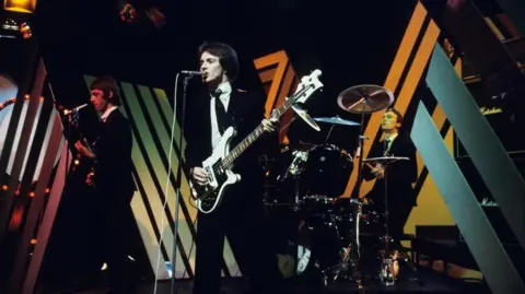 Rick Buckler is pictured on drums with The Jam bandmates Paul Weller (left) and Bruce Foxton (centre) performing in 1977 on the BBC's Top of the Pops show. They are dressed in black suits, white shirts and black ties against a BBC stage set with Marshall sound kit in the background. 