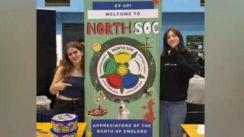 UoY Northern Society Two young women stand next to a banner promoting "North Soc" with a logo combining the Yorkshire and Lancashire roses.
