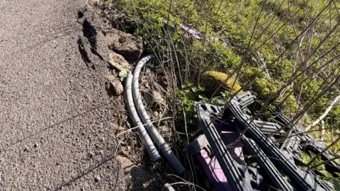 Cracked pavement and litter in the field next to it. The litter consists of plastic safety fencing and bits of pipe.