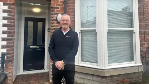 A man stands in front of a smart-looking, terraced, red-brick house and smiles towards the camera. He has cropped grey hair and wears a blue fleece over a white shirt, and black jeans. The house white a white bay window and a black door.