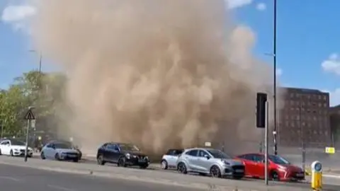 Adam Riding A huge cloud of brown dust which looks like a small tornado is seen billowing above a number of cars on a main road.