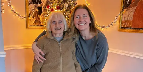 Two women standing close together, the younger woman on the right with her arm around the shorter older woman to our left. The woman on right is wearing a grey top and has brown shoulder length hair, while her mum has a beige zip-up fleece and grey hair in a bob. 