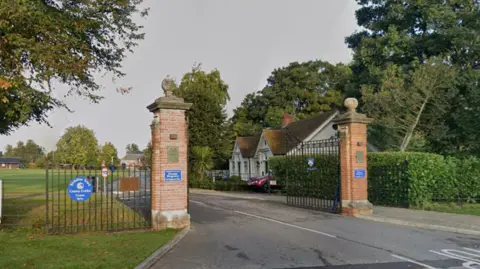 Gated entrance to Reading Blue Coat School. The tall brick pillars are topped with stone pediments. The grounds are parkland dotted with mature trees.