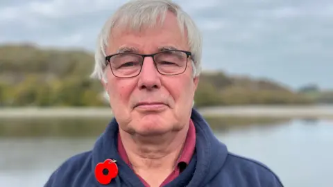 A man with grey hair and glasses, wearing a blue hoodie and a poppy stands in front of a bad of water, with a beach and dunes behind.