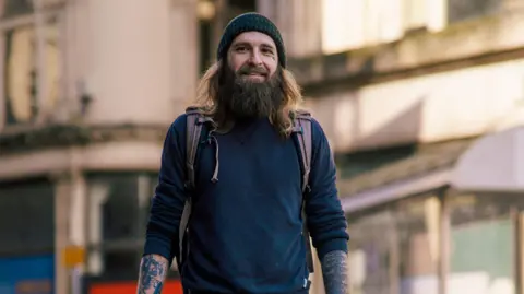 Tom Wilkins stood in Cardiff high street. He has a brown long beard, brown long hair and wears a knitted grey hat. Straps from his backpack can be seen on his shoulder, and he wears a jumper rolled up to his elbows 