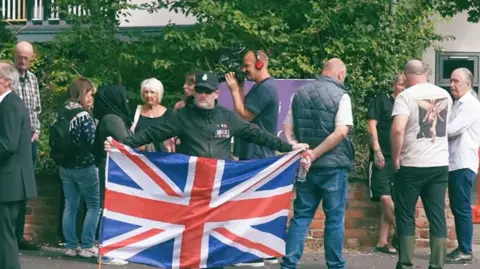 Protests outside the King's Gap hotel in Hoylake. A man is holding a Union Jack, and several other people standing around with the hotel in the background. 