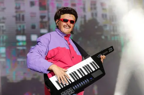 Picture of Oliver Tree in a purple and pink jacket performing in 2024. He is holding a keyboard.