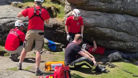 A group of people wearing helmets gather beside large rocky outcrops on a hillside, with rescue equipment spread out on the grass and rolling countryside in the background.