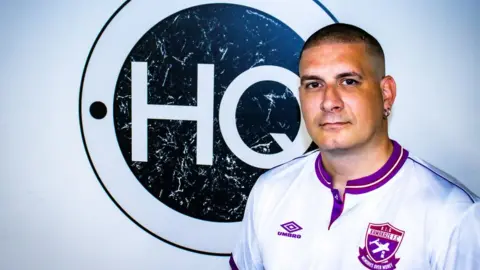 Yasin El Ashrafi Yasin El Ashrafi in a white football t-shirt in front of a HQ sign