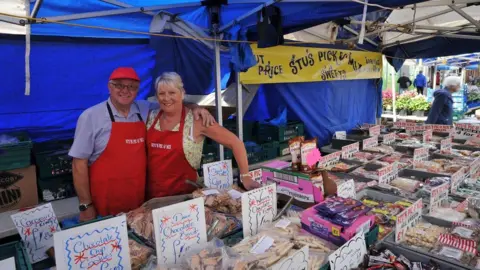 Stu and Bev Tooby stand behind their sweet stall at Nuneaton market. There is a blue canopy over the stall and many different types of products are laid out with signs. The couple are standing together, with their arms around each other.