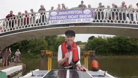 Tim Coghlan Lady Sheila is wearing a black cap, a red life jacket and a white top. She is holding a microphone while riding at the back of a boat which has just gone under a bridge with white railings. There are several people on the bridge, on which a sign saying BRAUNSTON HISTORIC BOAT SHOW is hanging.