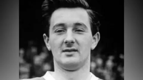 A black and white photograph of Jeff Hall, He is clean shaven and wears a v-neck shirt.