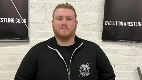 Photo of Liam Robinson, head coach at Evolution Pro Wrestling. He is wearing a black top and has a beard with his hair tied in a bun.