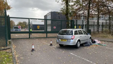 BBC/Sam Read Two people attached to a car outside the Lockheed martin gates