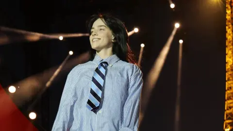 Getty Images Billie Eilish performs onstage during Global Citizen's Power Our Planet: Live in Paris on June 22, 2023 in Paris. Billie is a 22-year-old white woman with long black hair and green eyes. She wears a blue and white striped shirt with a blue and navy striped tie. She smiles, looking to the right of the camera. Behind her, spotlights can be seen against a dark sky and the Eiffel Tower, lit up, is just to the right of the frame