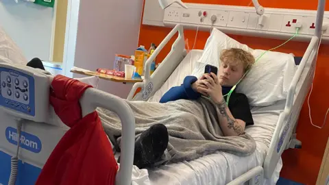 Supplied Harry Pyett, lying on a hospital bed while on his phone. His left arm is tattooed and he has a grey blanket over him.