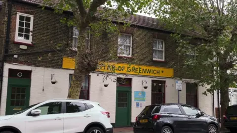 The exterior of a building "Star of Greenwich." It's a brick structure with a yellow sign above the entrance, bordered by trees on either side. Two cars are parked out front