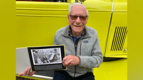 Justin Dealey/BBC Malcolm Stern holding a family photograph whilst sitting on the running board of the car