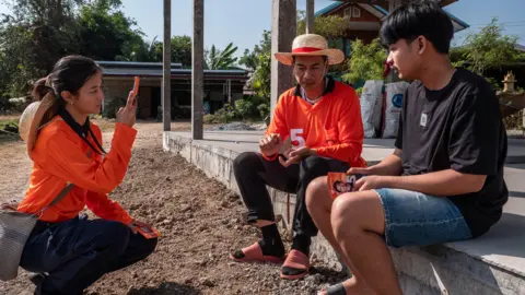 Jonathan Head/BBC Candidate Macky, centre, a man wearing an orange top and a hat, speaks to a young man wearing a black top while a colleague from his party (also in orange top) films on her mobile phone.