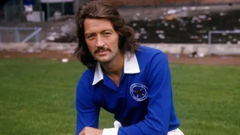 Frank Worthington is pictured during his playing days. He is crouched on a football pitch, wearing a long-sleeved royal blue football shirt with a white collar. He has a moustache and long dark brown hair.