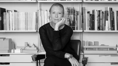 Valerie Bennett A black and white image of a woman in a striped top with one arm under her chin in front of a set of shelves with lots of books and magazines.