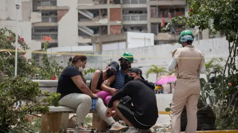A group of people console each other on a bench in front of a demolished building