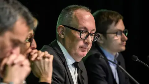 AFP via Getty Images Crans-Montana Mayor Nicolas Feraud looks downward as he sits with colleagues at a press conference where he revealed the ski bar had not been checked for five years. 