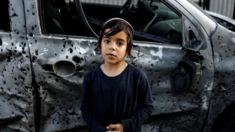 Reuters A boy stands next to a car that was damaged by rocket shrapnel in Sderot, Israel (2 May 2023)