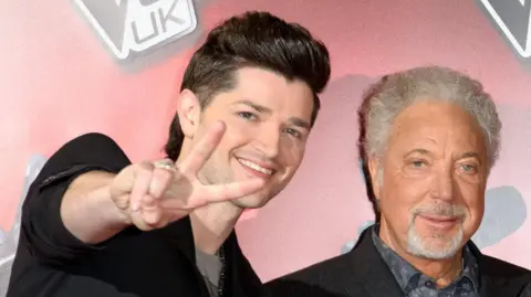 Danny O'Donoghue (left) and Sir Tom Jones (right) standing next to each other. They are both looking at the camera. Danny is doing a peace sign pose. They are both smiling.